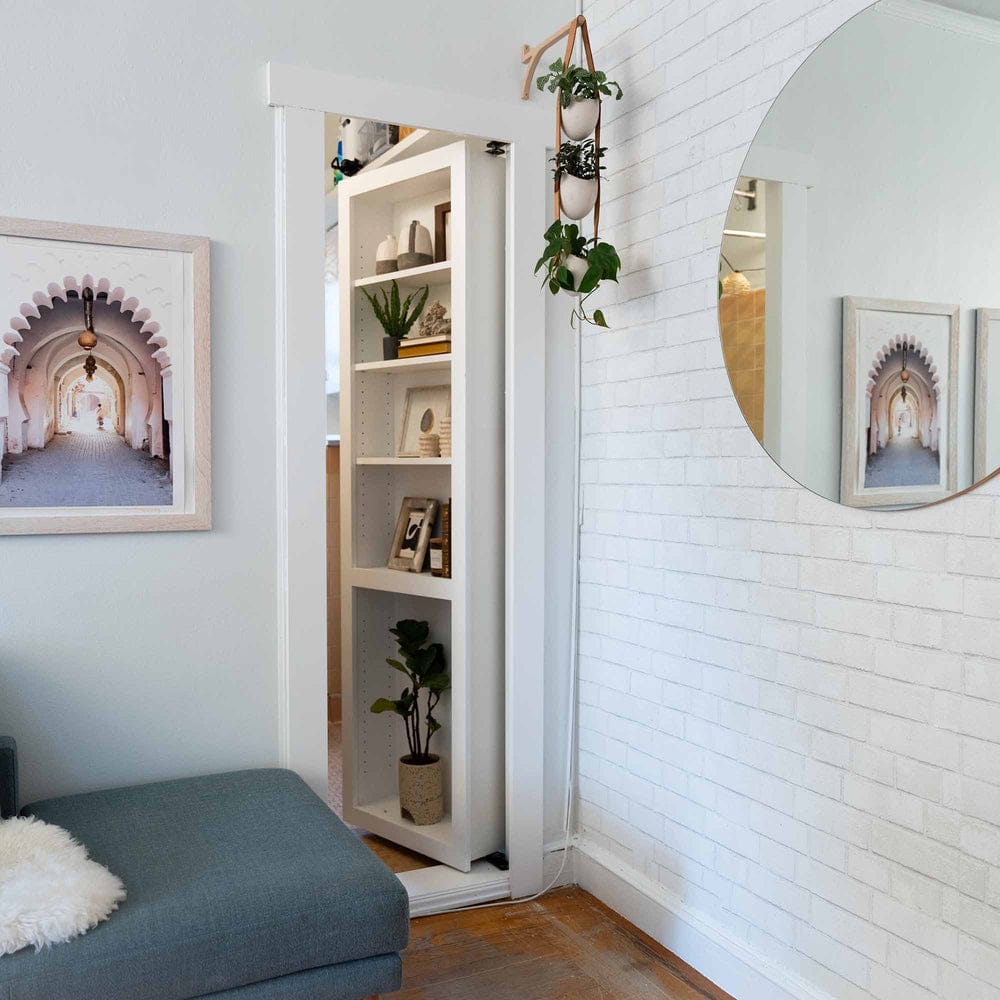 Murphy Door hidden bookcase door in white, closed, with shelves of plants and decor beside a blue couch